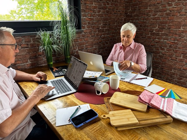 Zwei Personen arbeiten an einem Holztisch mit Laptops, Papieren und einem Smartphone. Im Hintergrund sind eine Ziegelwand und eine Pflanze zu sehen.